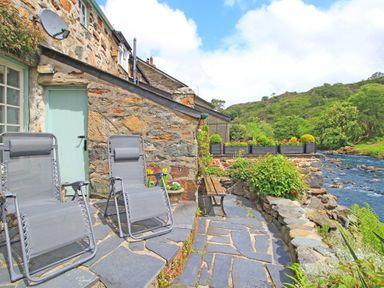A stone patio with two gray reclining chairs a bench and a river view at Ivy House in Beddgelert