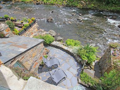 A stone patio with two lounge chairs and plants next to a flowing river at Ivy House in Beddgelert