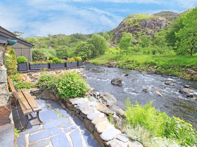 A stone patio with a bench and flower beds next to a river and green hills at Ivy House in Beddgelert