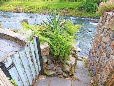 A stone pathway with steps leading down to a river with a wooden gate and plants on the side at Ivy House in Beddgelert