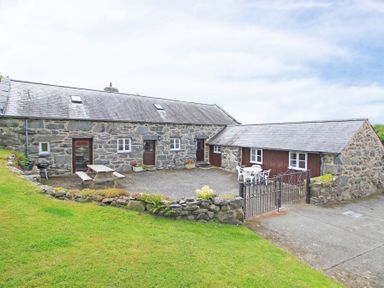 A stone cottage with two wooden doors two tables with chairs and a small stone wall at Y Beudy The Old Byre in Llanbedr