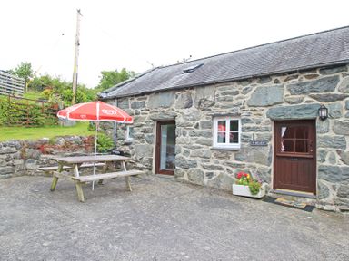 A stone cottage exterior with a picnic table and red umbrella on a concrete patio at Y Beudy The Old Byre in Llanbedr