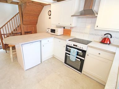 A kitchen with white cabinets a white microwave a red kettle a black oven and wooden chairs near stairs at Y Beudy The Old Byre in Llanbedr