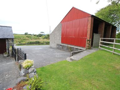 An outdoor area with a red barn next to a concrete driveway and a grassy lawn at Y Beudy The Old Byre in Llanbedr