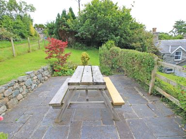 A wooden picnic table on a stone patio with a garden and greenery surrounding it at Y Beudy The Old Byre in Llanbedr