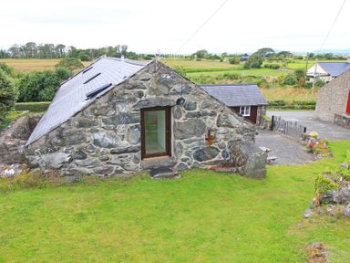 A stone house with a slate roof surrounded by green grass and fields at Y Beudy The Old Byre in Llanbedr