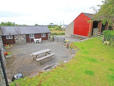 A courtyard with a wooden picnic table and white plastic chairs next to a stone and wood building and a red barn at Y Beudy The Old Byre in Llanbedr