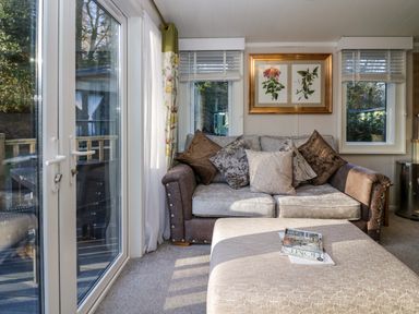 A small sitting area with a sofa, cushions, ottoman, framed botanical prints, windows with blinds, and glass doors leading outside at Lily Rose Lodge in White Cross Bay near Troutbeck Bridge
