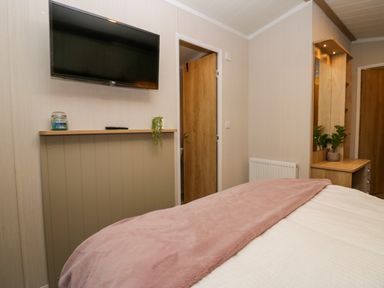 A bedroom with a bed covered with a pink blanket a wall-mounted television and a wooden vanity table with a mirror at Lily Rose Lodge White Cross Bay near Troutbeck Bridge