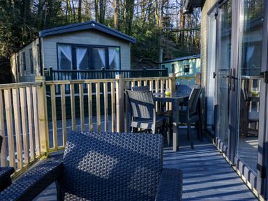 An outdoor deck with woven chairs and a dining table with a candle near holiday lodges in a wooded area at Lily Rose Lodge White Cross Bay near Troutbeck Bridge