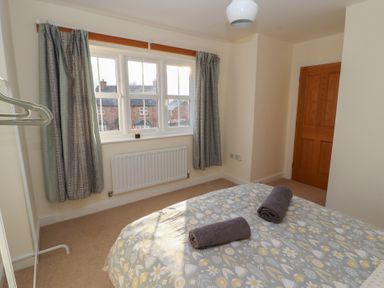 A bedroom with a bed covered in a patterned duvet and two towels on top near a window with curtains and a wooden door at 4 Centurion Rise in Penrith
