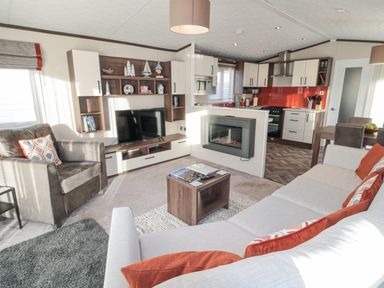A living room with a sofa and armchair a TV mounted on a wooden wall unit a coffee table a fireplace and a kitchen with dining area at Downtime Lodge near Felton