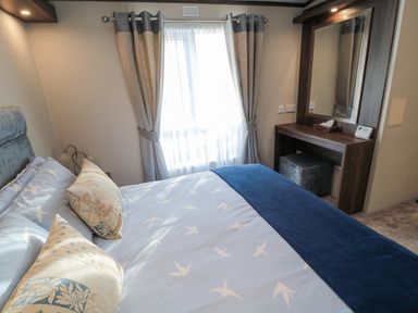 A bedroom with a bed covered with patterned bedding and two pillows a window with curtains and a wooden dressing table with a mirror and a stool at Downtime Lodge near Felton