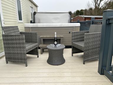 An outdoor deck with two wicker chairs a wicker table holding a bottle and two glasses in front of a hot tub