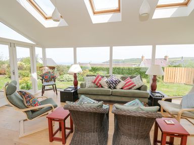 A conservatory with a green sofa pillows wicker chairs wooden rocking chair and large windows at Tree Tops in Newport Pembrokeshire