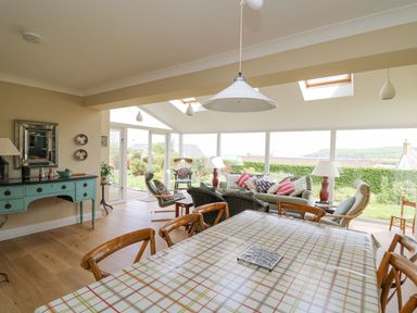 A dining area with a checkered tablecloth and wooden chairs next to a seating area with sofas and armchairs by large windows at Tree Tops in Newport Pembrokeshire
