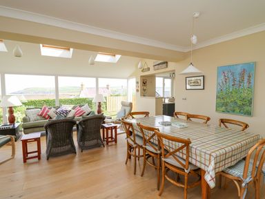 A dining area with a checkered tablecloth and wooden chairs next to a living room with wicker chairs and a sofa at Tree Tops in Newport Pembrokeshire