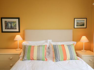 A bedroom with a bed and two striped pillows two bedside tables with lamps and framed pictures on a yellow wall at Tree Tops in Newport Pembrokeshire