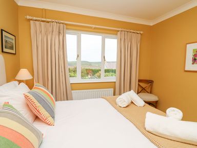 A bedroom with a bed, striped pillows, rolled white towels, a lamp, a chair, a window with beige curtains, and a framed picture at Tree Tops in Newport Pembrokeshire