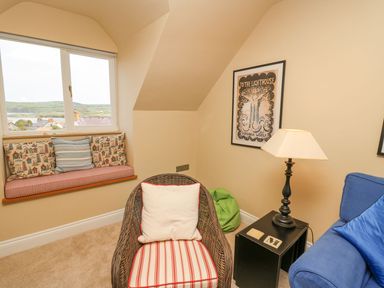 A sitting area with a window seat and cushions a wicker chair with a cushion a blue sofa a side table with a lamp and framed artwork on the wall at Tree Tops in Newport Pembrokeshire
