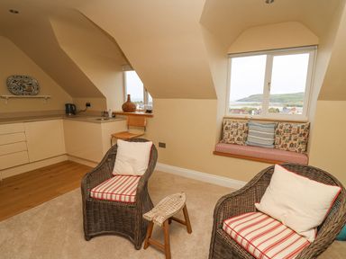 A small kitchen area with cream cabinets and a kettle a wicker chair with a cushion a window seat with cushions and a small decorative wooden stool at Tree Tops in Newport Pembrokeshire
