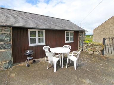 An outdoor patio with a round white table and four white plastic chairs beside a wooden building with two windows at Y Stabl The Old Stable in Llanbedr