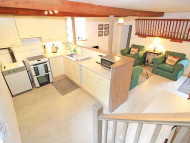 A kitchen with white cabinets stove dishwasher and microwave next to a sitting area with green armchairs and a lamp at Y Stabl The Old Stable in Llanbedr