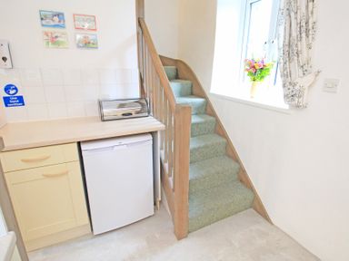 A kitchen corner with a small fridge under the counter and stairs next to a window with flowers in a vase at Y Stabl The Old Stable in Llanbedr