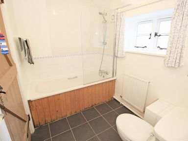 A bathroom with a bathtub that has a wooden panel and shower screen a white toilet a window with curtains and a radiator below at Y Stabl The Old Stable in Llanbedr