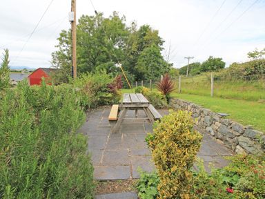 An outdoor patio area with a wooden picnic table on stone tiles surrounded by plants and a stone wall at Y Stabl The Old Stable in Llanbedr