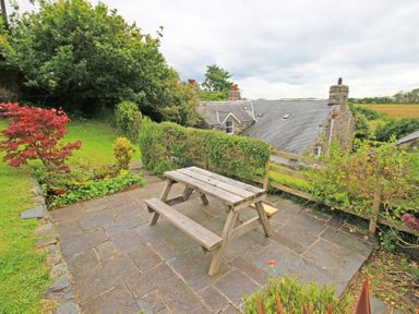 An outdoor patio area with a wooden picnic table surrounded by greenery and stone walls at Y Stabl in Llanbedr