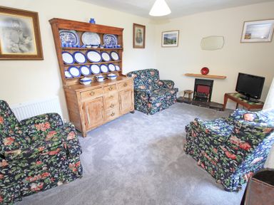 A living room with floral armchairs a wooden cabinet with blue and white plates a small fireplace and a television at The Farmhouse in Llanbedr