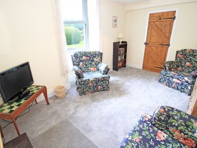 A sitting room with floral armchairs a wooden door a television on a table and a small bookshelf at The Farmhouse in Llanbedr