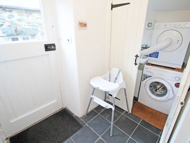 A utility room with a washing machine and dryer stacked in a cupboard and a white high chair on a tiled floor at The Farmhouse in Llanbedr