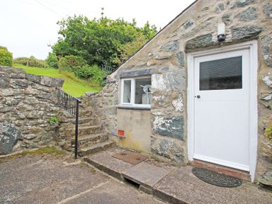 Stone exterior wall with a white door and window and stone steps with metal railing leading to grass at The Farmhouse in Llanbedr