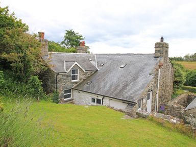 A stone cottage with a slate roof and chimneys surrounded by grass and trees at The Farmhouse in Llanbedr