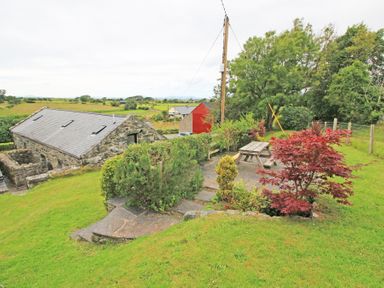 A stone building with skylights next to a grassy area with bushes trees and a wooden picnic table at The Farmhouse in Llanbedr