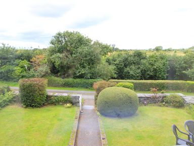 A garden with trimmed bushes and a pathway leading to a road with trees and hedges in the background at The Farmhouse in Llanbedr