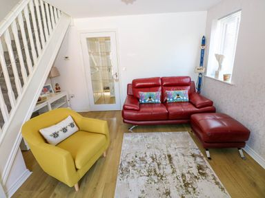 A living room with a yellow armchair and a red sofa with two cushions a footstool a staircase and wooden floor at Ty Bach Twt Llanelli