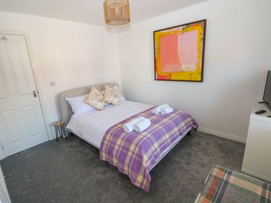 A bedroom with a double bed with purple plaid blanket towels and floral pillows a colorful abstract painting on the wall at Ty Bach Twt Llanelli