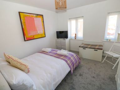 A bedroom with a bed with a purple blanket and two rolled towels a white dresser with a TV a plaid bench a white folding chair and two windows at Ty Bach Twt Llanelli