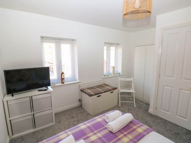 A bedroom with a bed covered in a purple plaid blanket two rolled towels a TV on a storage unit a white chair and a storage bench at Ty Bach Twt Llanelli