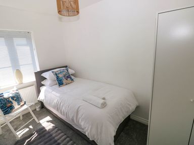 A small bedroom with a single bed white bedding pillows chair with cushions window and a wardrobe at Ty Bach Twt Llanelli