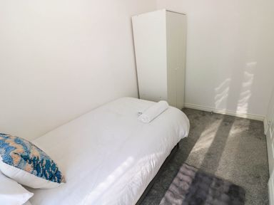 A small bedroom with a single bed white bedding a patterned pillow a rolled towel a white wardrobe and grey carpet at Ty Bach Twt Llanelli