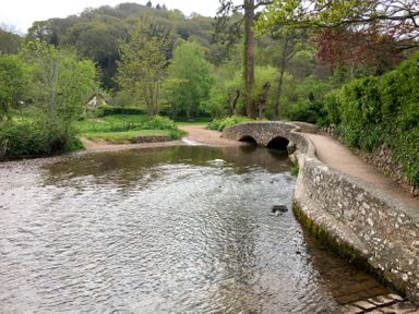 A stone bridge over a shallow river with a dirt path and trees at Little Dragons in Dunster near Minehead