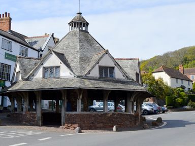 A stone and wood market building with a shingled roof and small windows in a village setting at Little Dragons in Dunster near Minehead