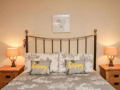 A bedroom with a metal bed frame, patterned bedding, two pillows with happy text, wooden bedside tables, lamps, and a picture on the wall at Little Dragons in Dunster near Minehead