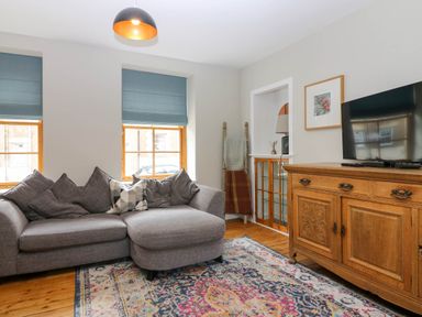 A living room with a gray sectional sofa and patterned rug a wooden cabinet with a television on top and two windows with blue blinds at 49 Castlegate in Jedburgh