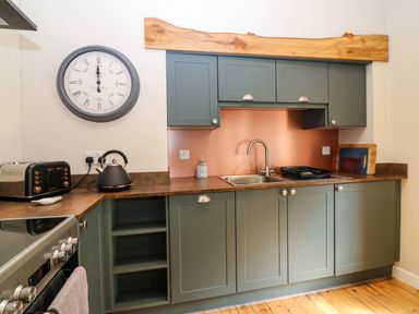A kitchen with green cabinets a metal sink wooden cutting boards a black toaster and kettle and a wall clock at 49 Castlegate in Jedburgh