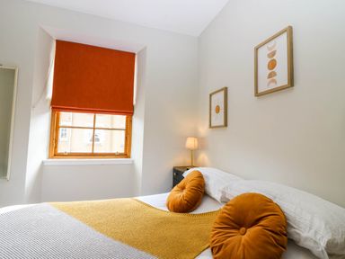 A bedroom with orange cushions a yellow throw a window with an orange blind and framed wall art at 49 Castlegate in Jedburgh